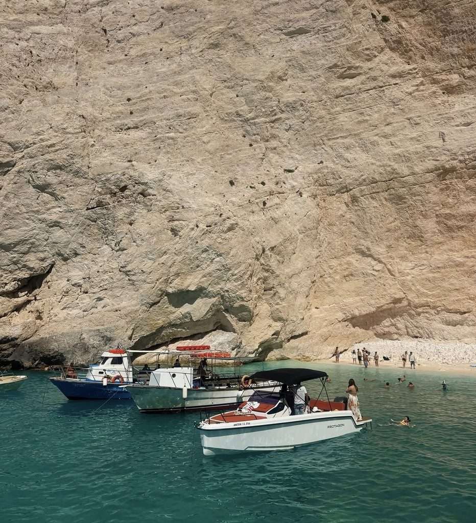 White beach at Shipwreck Beach in Zante, with boats resting on the turquoise water beneath the towering cliffs during a Zante shipwreck boat tour.